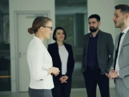 Geschäftsleute sprechen miteinander in einem offenen, modernen Büroflur mit Glaswänden und hellen Räumen.