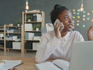 Junge Frau sitzt am Schreibtisch, spricht am Telefon und nutzt einen Laptop für Büroarbeit