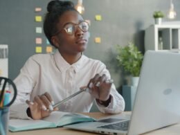 Junge Frau mit Brille sitzt an einem Schreibtisch und arbeitet am Laptop in einem modernen Büro
