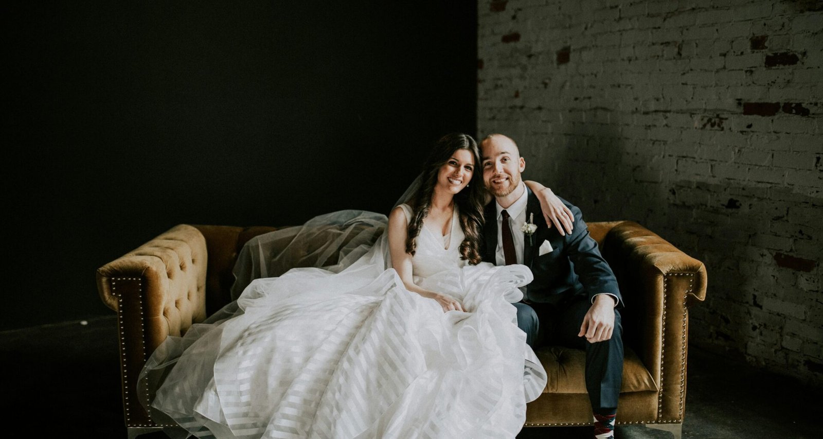 Lächelnder Bräutigam und lächelnde Braut im Hochzeitsoutfit sitzen gemeinsam auf einem Sofa