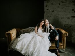 Lächelnder Bräutigam und lächelnde Braut im Hochzeitsoutfit sitzen gemeinsam auf einem Sofa