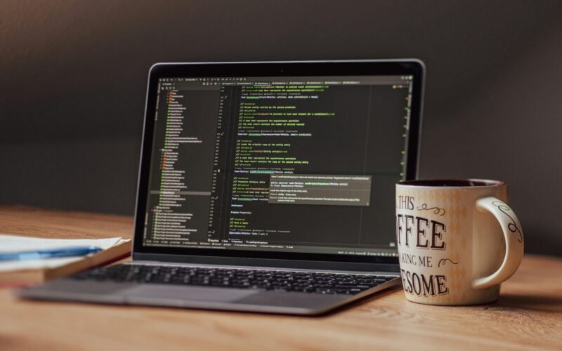 Offener Laptop mit angezeigtem Programmiercode, daneben eine weiße Kaffeetasse auf einem Tisch