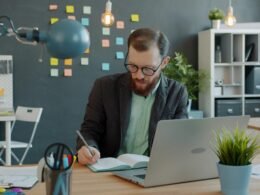 Mann arbeitet an einem Schreibtisch mit Laptop und Notizbuch in einem modernen Büro