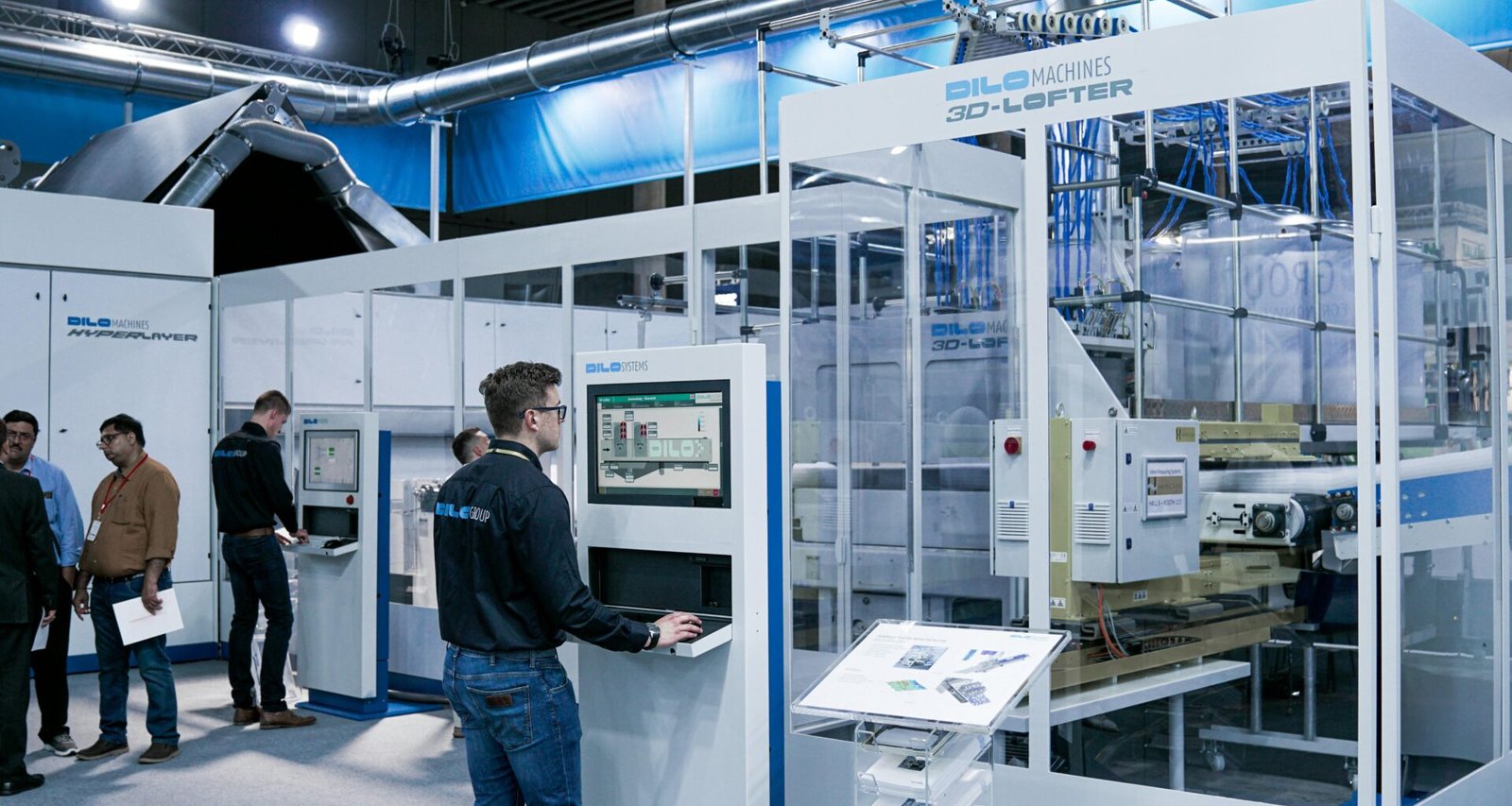 Ein Mann steht am Computer, während im Hintergrund einige Kollegen miteinander sprechen – typische Szene im modernen Büro