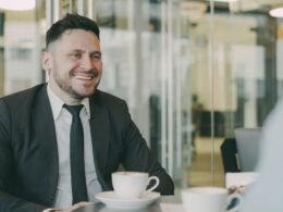 Ein Mann im Anzug sitzt lächelnd mit einer Tasse Kaffee an einem Tisch in einem modernen Café.