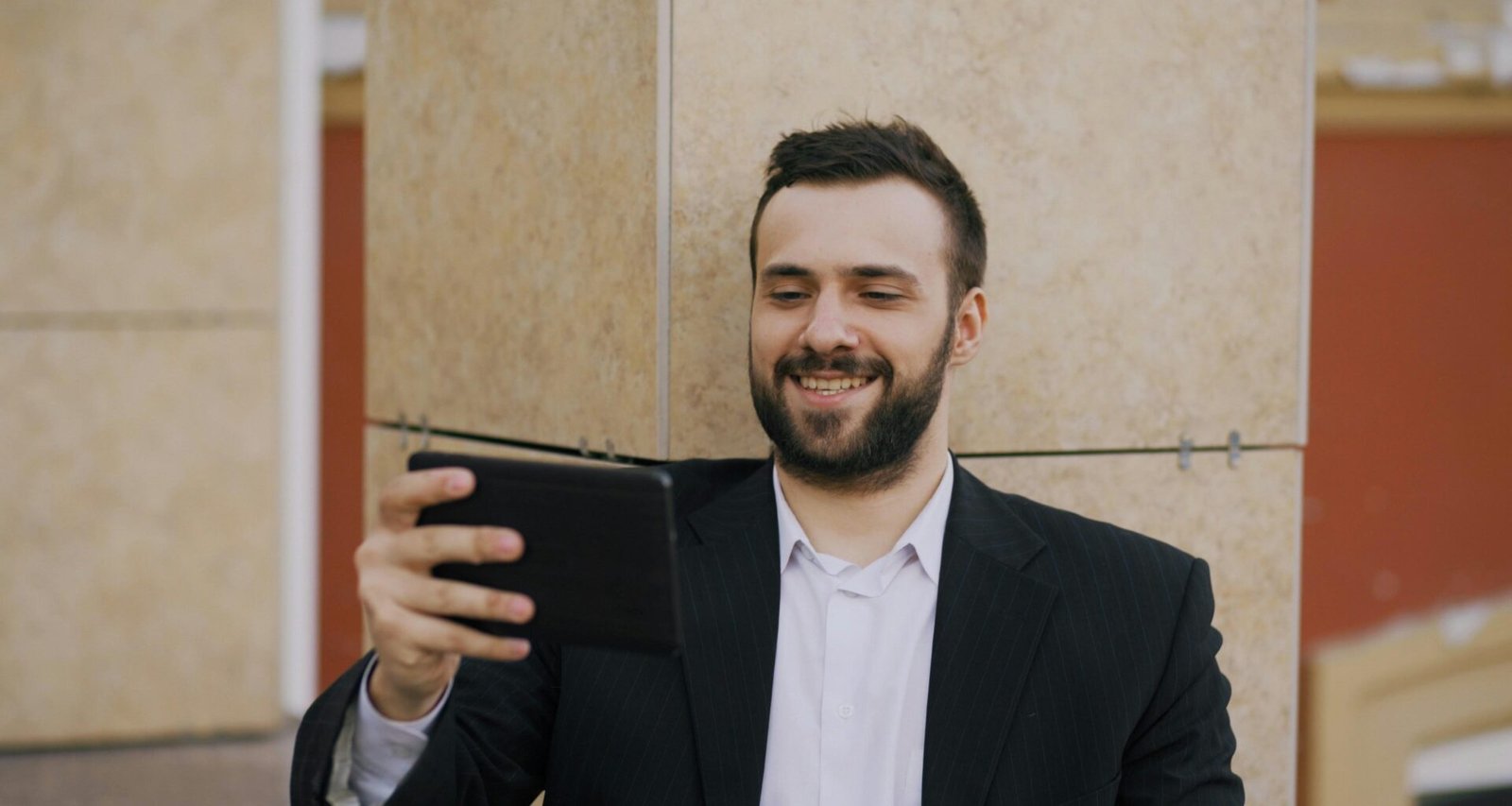 Ein Mann im Anzug sitzt und lächelt, während er auf ein Tablet blickt, in einem modernen Büro.