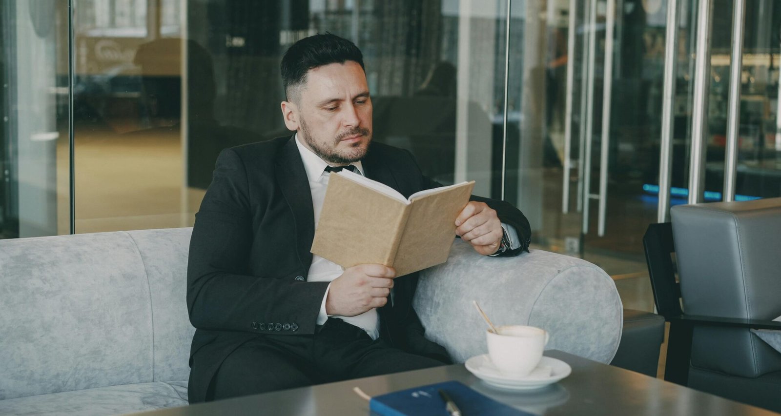 Mann im Anzug sitzt an einem Tisch, liest ein Buch und trinkt Kaffee in einem modernen Büro