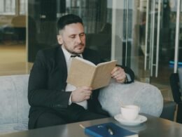 Mann im Anzug sitzt an einem Tisch, liest ein Buch und trinkt Kaffee in einem modernen Büro