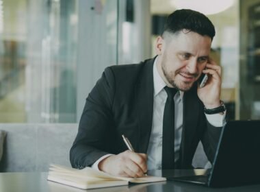 Ein Mann im Anzug spricht am Telefon und schreibt in ein Notizbuch an einem Schreibtisch im Büro.