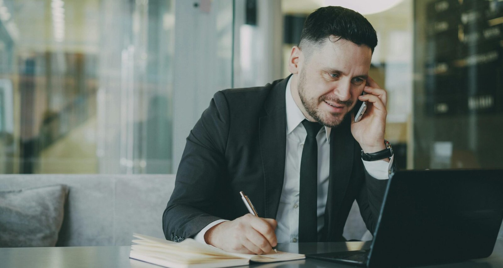 Mann im Business-Anzug telefoniert und schreibt währenddessen in ein Notizbuch an einem Schreibtisch