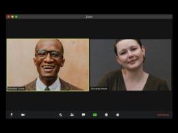 Lächelnder Mann in schwarzem Anzug sitzt frontal und blickt in die Kamera vor neutralem Hintergrund