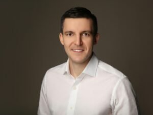 Mann in weißem Hemd steht lächelnd vor neutral grauem Hintergrund, modernes Porträtfoto für Business-Artikel