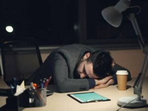 Mann schläft am Schreibtisch neben einer Kaffeetasse und einem Tablet, symbolisch für Müdigkeit im Arbeitsalltag.