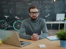 Ein Mann arbeitet sitzend an einem Schreibtisch vor einem Laptop, hinter ihm steht ein Fahrrad an der Wand.