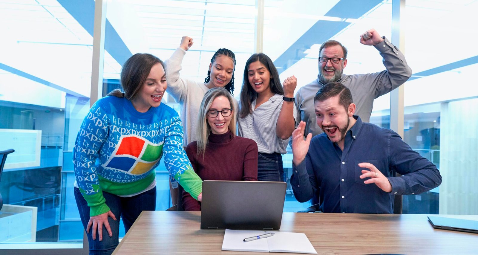 Mehrere Männer und Frauen sitzen und stehen um einen Tisch und schauen gemeinsam auf einen Laptop während eines Meetings.