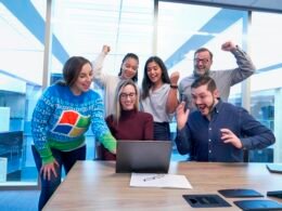 Mehrere Männer und Frauen sitzen und stehen um einen Tisch und schauen gemeinsam auf einen Laptop während eines Meetings.