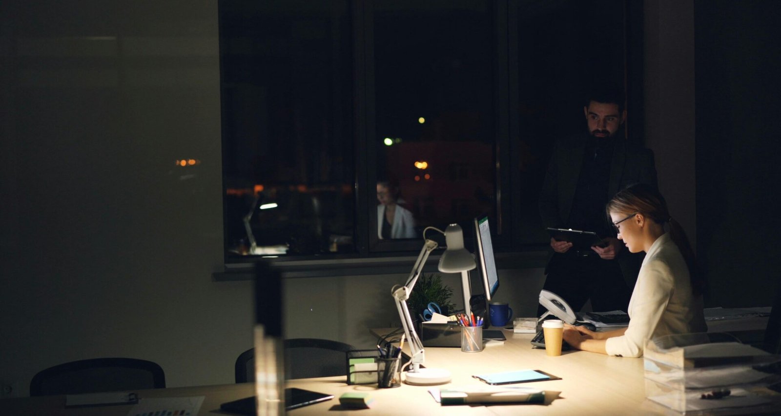 Personen arbeiten in einem gedimmten Büro bei Nacht an ihren Computern und sind auf Bildschirme fokussiert.