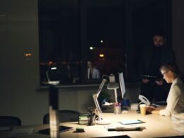 Personen arbeiten in einem gedimmten Büro bei Nacht an ihren Computern und sind auf Bildschirme fokussiert.
