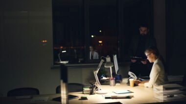 Personen arbeiten in einem gedimmten Büro bei Nacht an ihren Computern und sind auf Bildschirme fokussiert.
