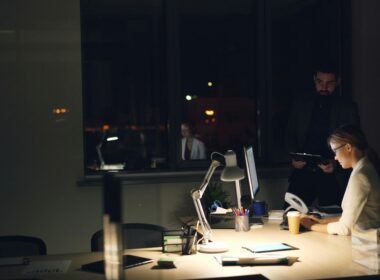 Personen arbeiten in einem gedimmten Büro bei Nacht an ihren Computern und sind auf Bildschirme fokussiert.