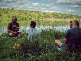 Mehrere Personen befinden sich am Ufer eines Sees, umgeben von natürlicher Landschaft und Wasserfläche