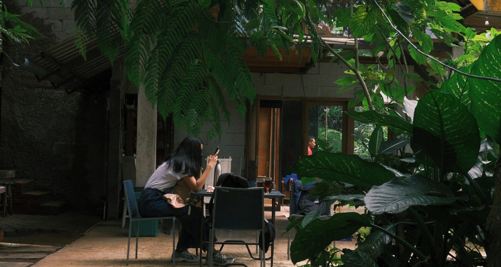 Menschen sitzen an mehreren Tischen in einem von vielen Pflanzen beschatteten Open-Air-Café und unterhalten sich.