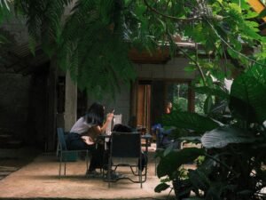 Menschen sitzen an mehreren Tischen in einem von vielen Pflanzen beschatteten Open-Air-Café und unterhalten sich.