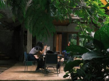 Menschen sitzen an mehreren Tischen in einem von vielen Pflanzen beschatteten Open-Air-Café und unterhalten sich.
