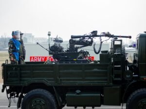 Ein Militär-LKW mit aufgesetzter Waffe, begleitet von mehreren Soldaten während eines Einsatzes auf unbefestigtem Gelände.