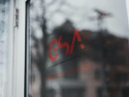 Nahaufnahme eines Fensters, dahinter ist ein moderner Gebäudekomplex im Hintergrund unscharf zu erkennen.
