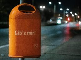 Orangefarbener Metall-Mülleimer steht bei Nacht am Straßenrand unter künstlicher Beleuchtung.