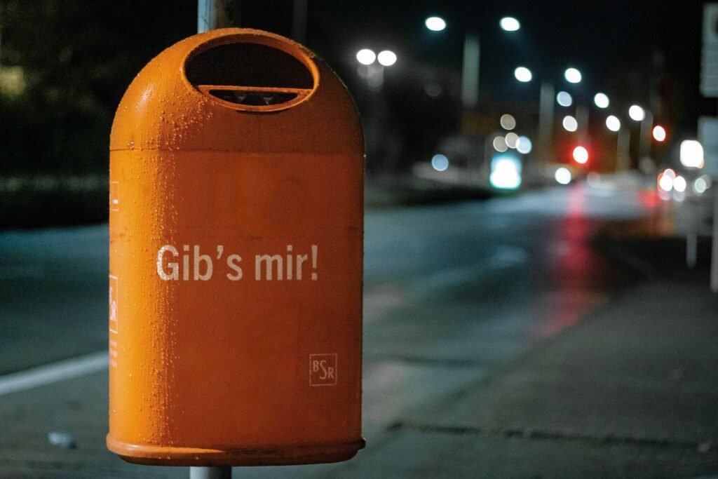 Orange Mülltonne aus Metall am Straßenrand bei Nacht, beleuchtet von Straßenlaternen, städtisches Umfeld