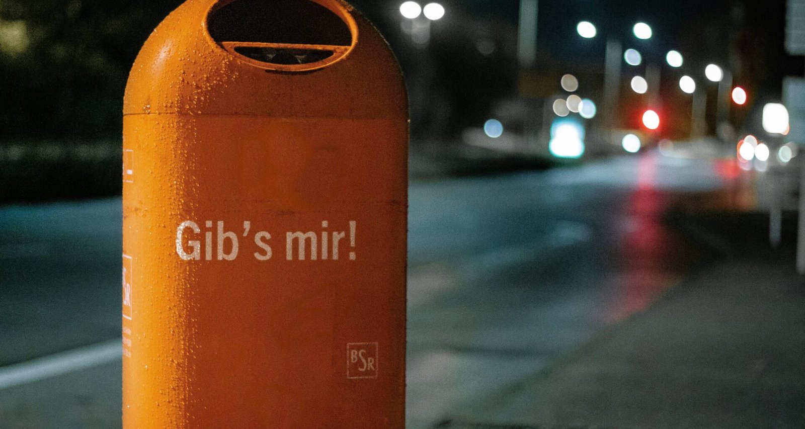 Orange Mülltonne aus Metall am Straßenrand bei Nacht, beleuchtet von Straßenlaternen, städtisches Umfeld
