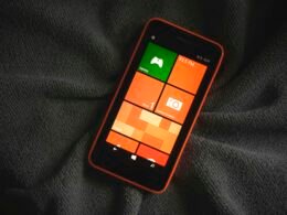 Orangefarbenes Smartphone mit bunten Kacheln auf dem Bildschirm, das auf einem neutralen Hintergrund liegt