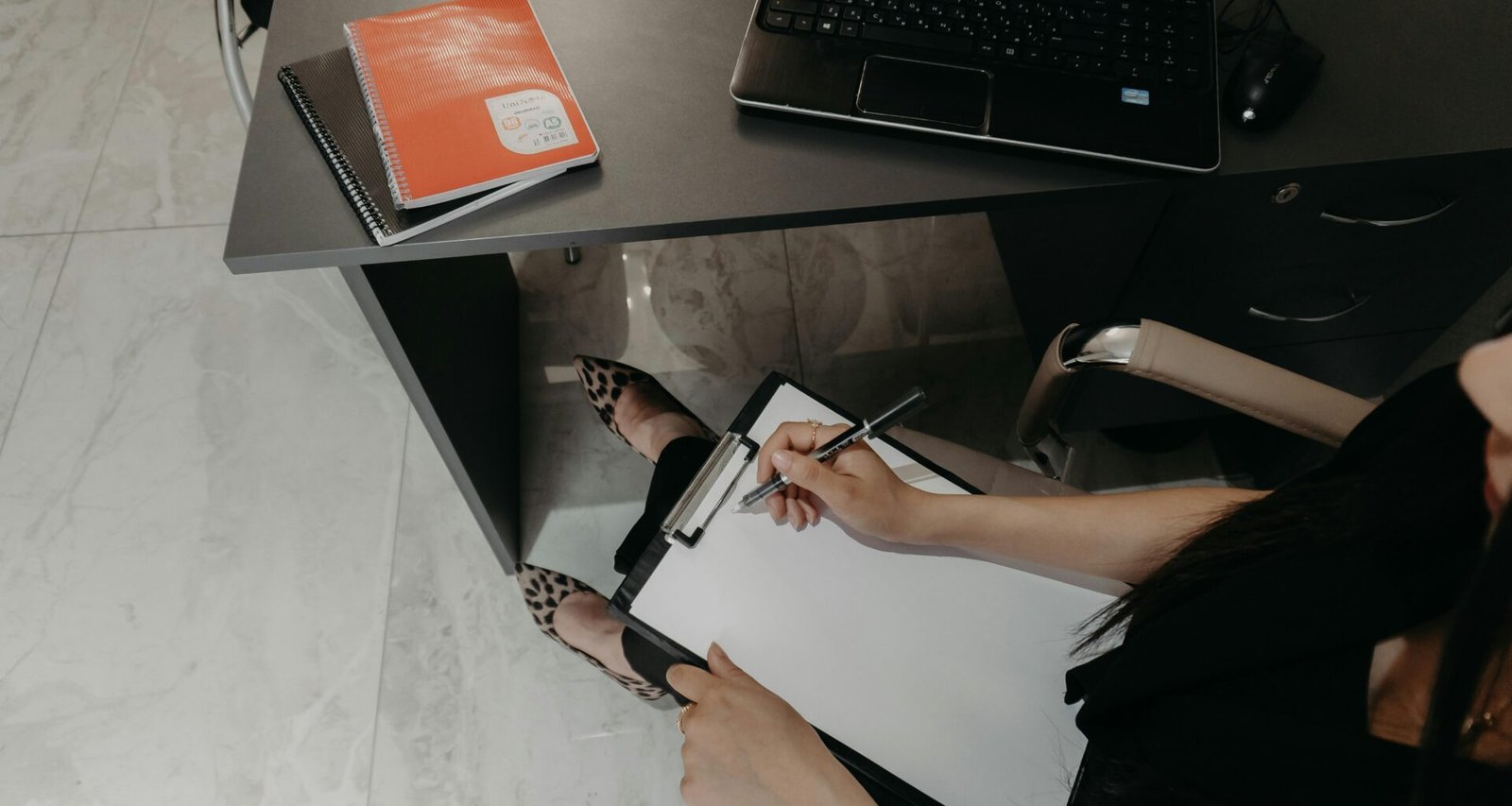 Person schreibt Notizen auf einem Klemmbrett am Schreibtisch mit offenem Laptop, Fokus auf digitale Arbeitsweise