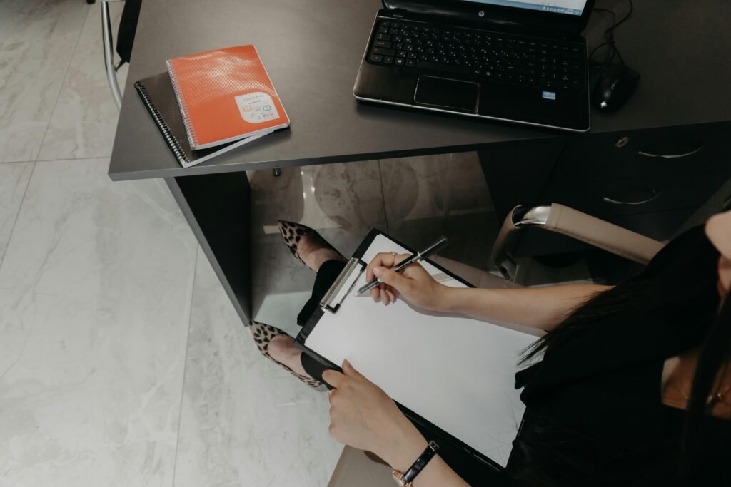 Person sitzt am Schreibtisch, schreibt auf ein Klemmbrett und arbeitet mit einem Laptop, ideal für Artikel zu Büroorganisation