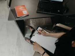 Person sitzt am Schreibtisch, schreibt auf ein Klemmbrett und arbeitet mit einem Laptop, ideal für Artikel zu Büroorganisation