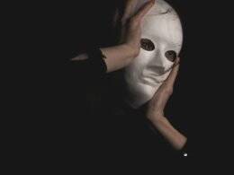 Person mit schwarzem Langarmshirt sitzt und hält die Hände vors Gesicht, neutraler Hintergrund