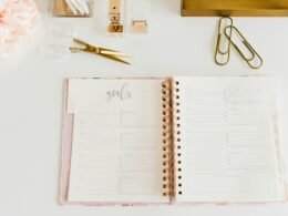 Offenes Spiralnotizbuch, daneben eine Schere und zwei Büroklammern auf einem weißen Tisch. Bürobedarf zur Organisation.