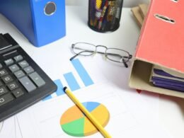 Schreibtisch mit Taschenrechner, ausgedruckten Diagrammen und mehreren Ordnern, geeignet für Themen rund um Büroarbeit und Finanzen.