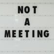 Schwarzes Schild mit der Aufschrift 'Note a meeting' an einer weißen Wand in einem modernen Bürogebäude.
