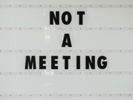 Schwarzes Schild mit der Aufschrift 'Note a meeting' an einer weißen Wand in einem modernen Bürogebäude.