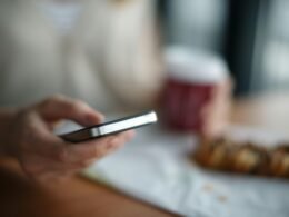 Person hält ein graues Smartphone in der Hand, im Vordergrund fokussiert; unscharfer Hintergrund unterstreicht den Fokus aufs Gerät.