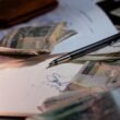Geldscheine und ein Kugelschreiber liegen auf mehreren Dokumenten als Symbol für Finanzplanung und Budgetierung.