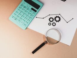 Ein Taschenrechner, eine Lupe und ein Papier mit Diagramm und Zahnrädern zeigen Analyse- und Optimierungsprozesse im Online-Marketing.