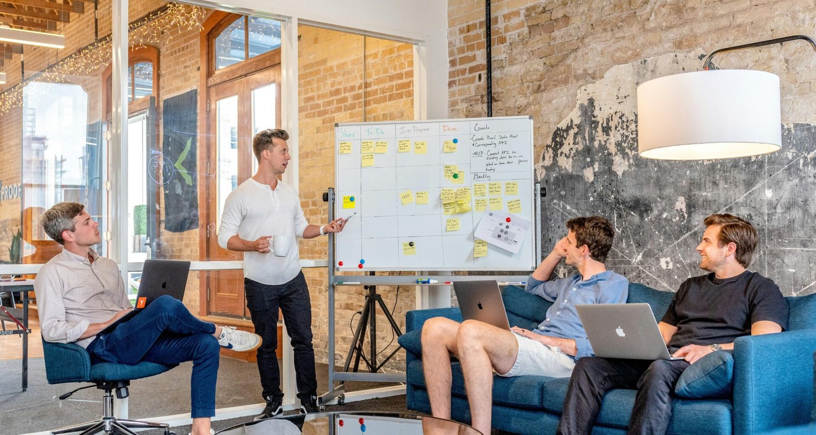 Drei Männer sitzen mit Laptops und verfolgen einen Mann, der neben einem Whiteboard präsentiert