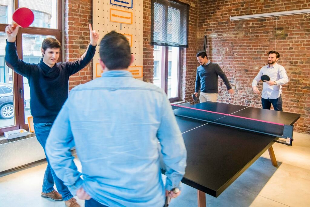 Vier Männer spielen Tischtennis in einem modernen Büro mit Glaswänden, umgeben von Arbeitsplätzen und entspannter Atmosphäre.