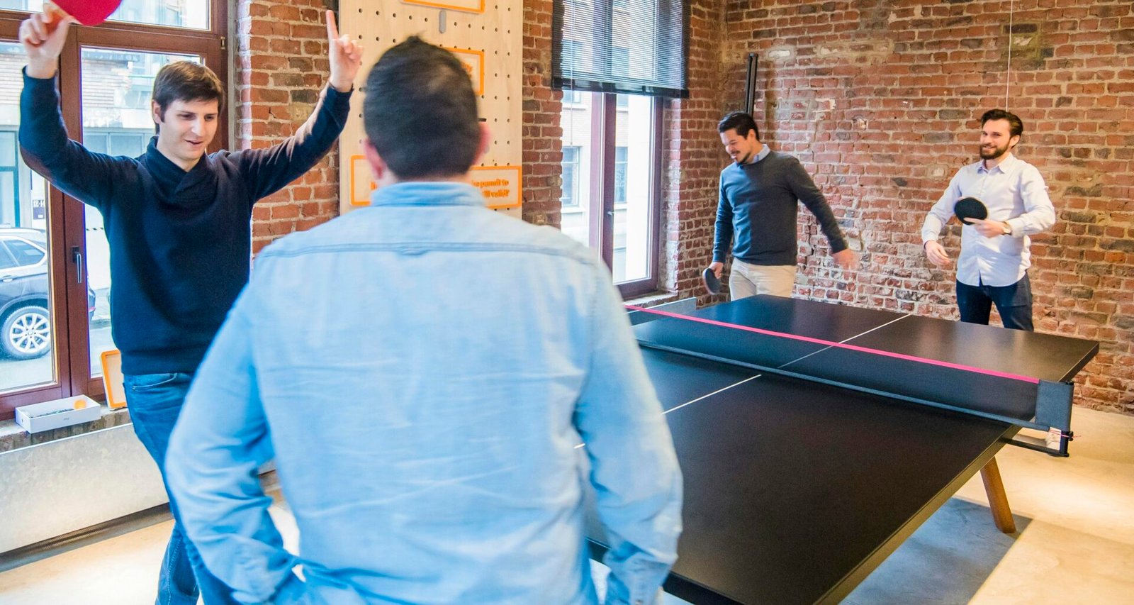 Vier Männer spielen Tischtennis in einem modernen Büro mit Glaswänden, umgeben von Arbeitsplätzen und entspannter Atmosphäre.