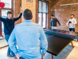 Vier Männer spielen Tischtennis in einem modernen Büro mit Glaswänden, umgeben von Arbeitsplätzen und entspannter Atmosphäre.