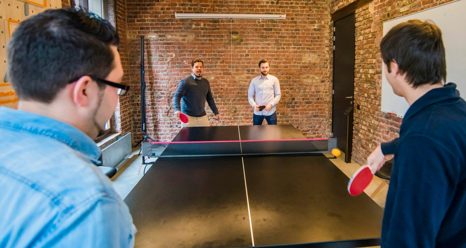 Vier erwachsene Männer spielen gemeinsam Tischtennis in einem modernen Büro mit großen Fenstern und Tageslicht.